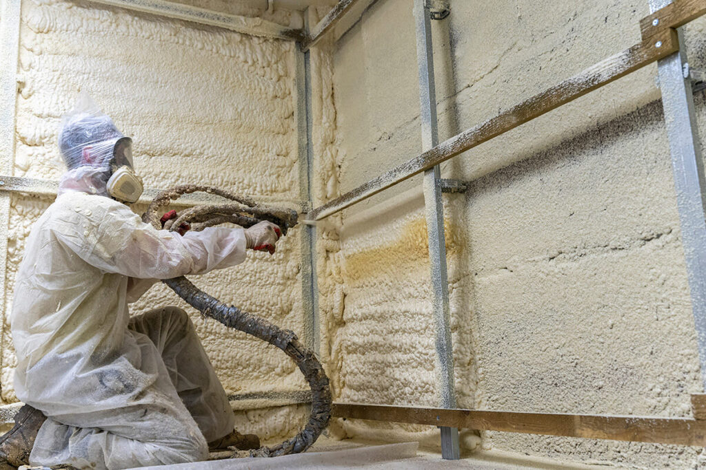Worker applying spray foam insulation on wall and pipes.
