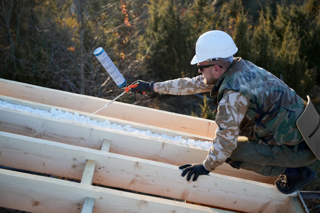 Construction worker in safety helmet applying sealant on wooden structure outdoors.
