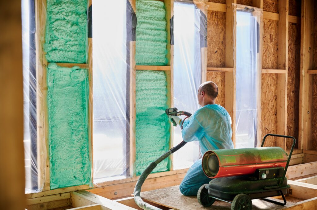 Person spraying green insulation foam inside a wooden frame wall.